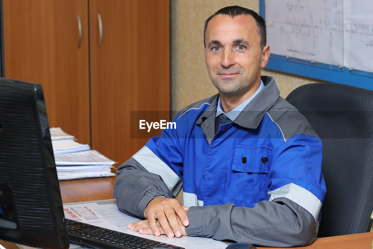 A young man in overalls is sitting at a desk in a chair. a cheerful engineer of caucasian appearance 