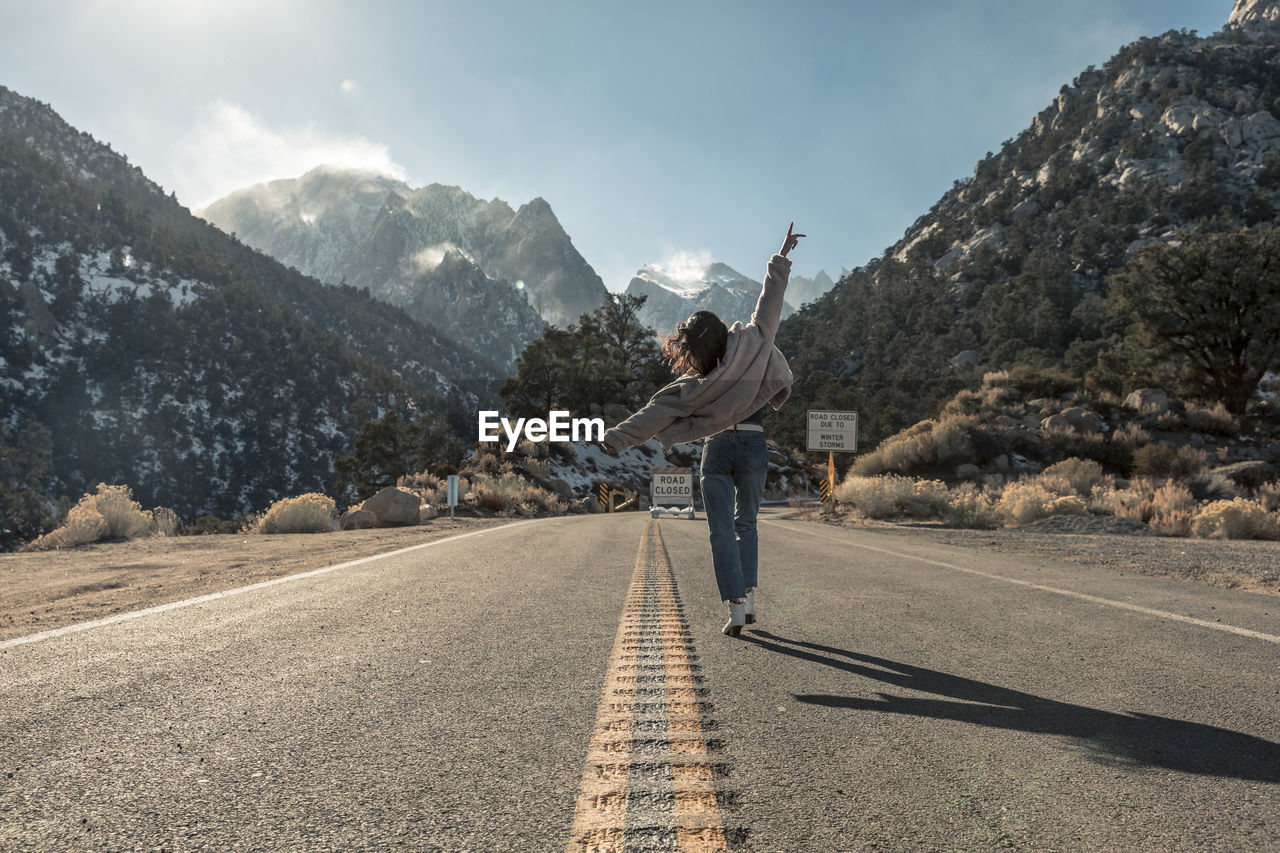 Full length of young woman wearing warm clothing while walking on road against mountains during sunny day