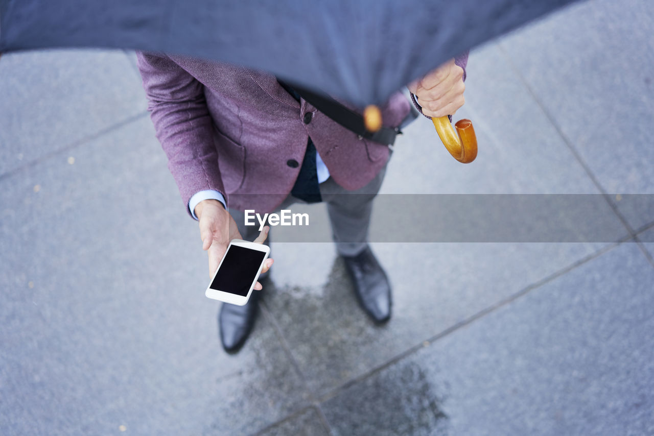 Businessman standing under umbrella holding cell phone, partial view