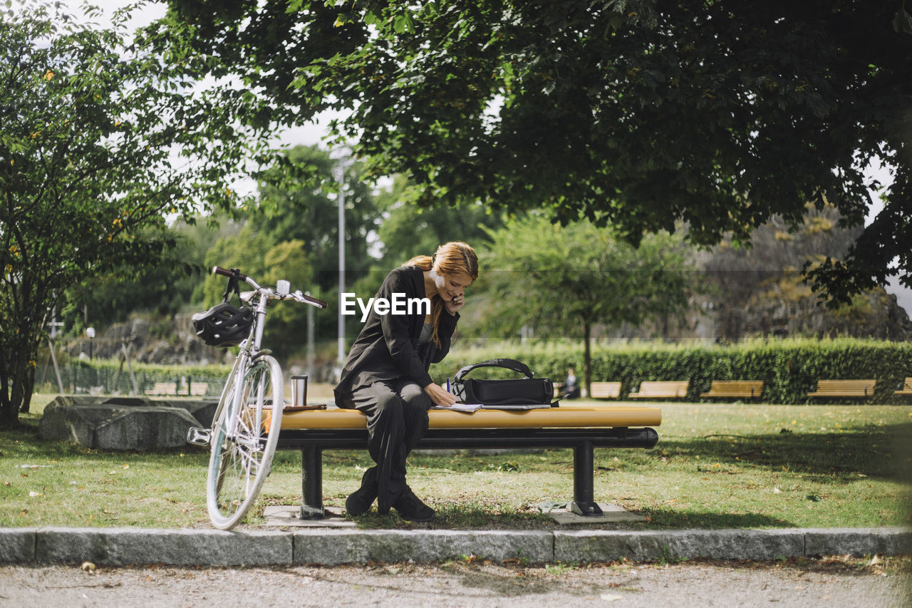 Full length of businesswoman talking on smart phone while sitting by bicycle at park