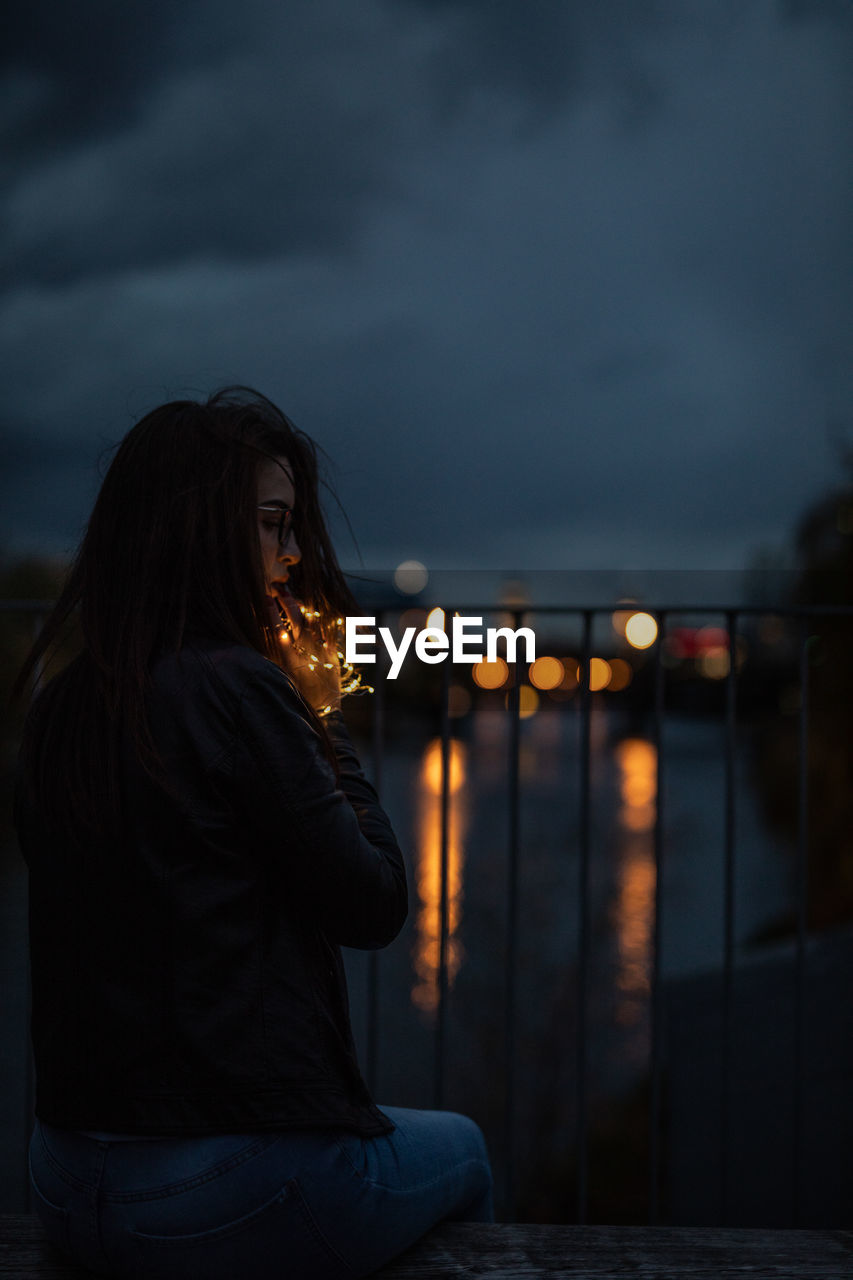 Woman sitting with illuminated string lights on railing against river at night