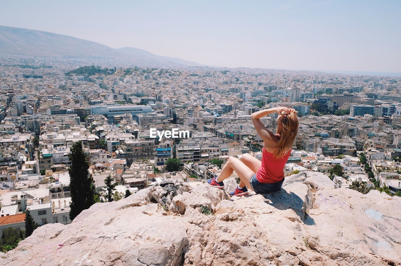 Rear view of woman sitting on rock formation while looking at city against sky