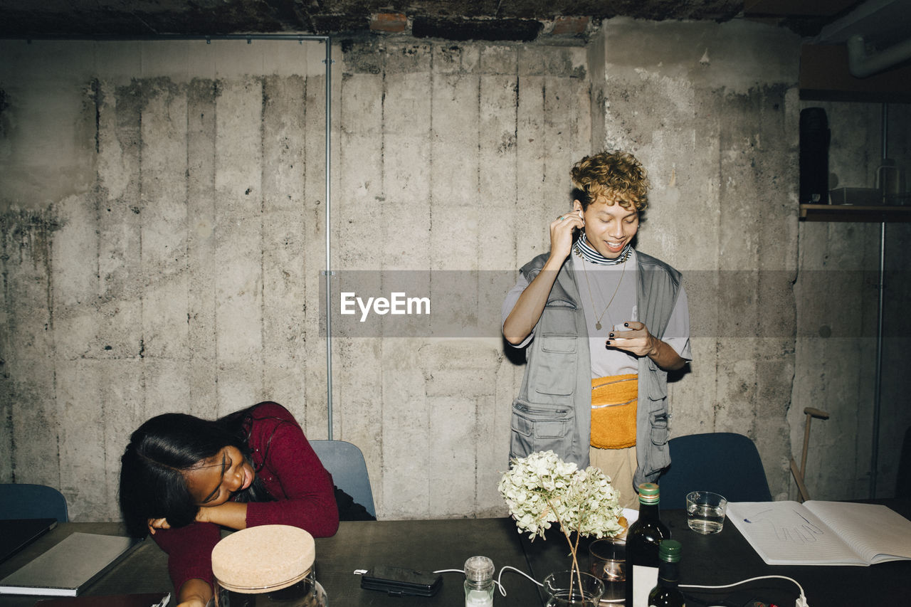 Smiling entrepreneur wearing in-ear headphones while female coworker sleeping at table in office