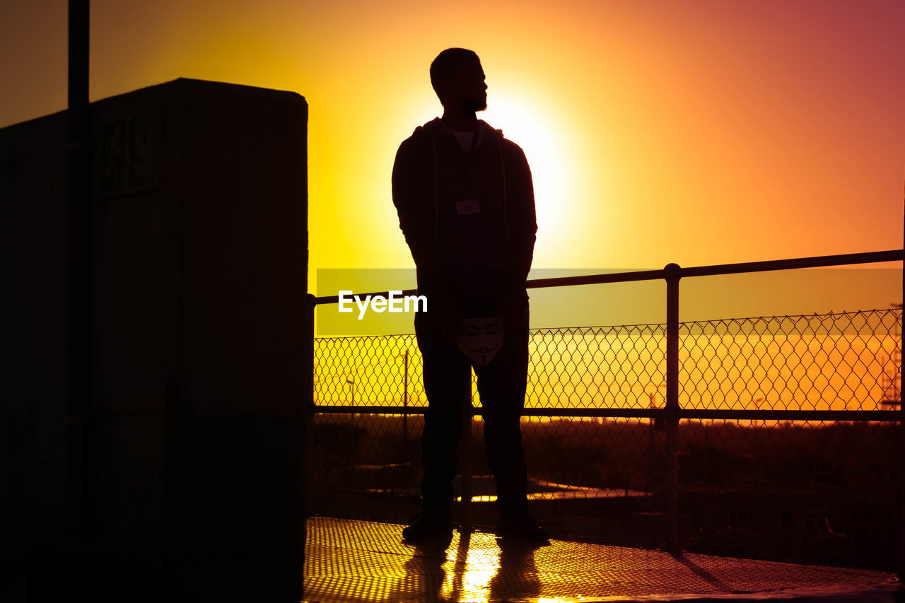 Low angle view of silhouette man standing by chainlink fence against sky during sunset