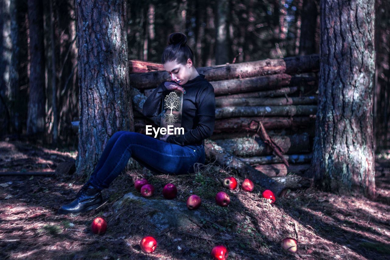 Full length of young woman holding decoration while sitting by apples in forest