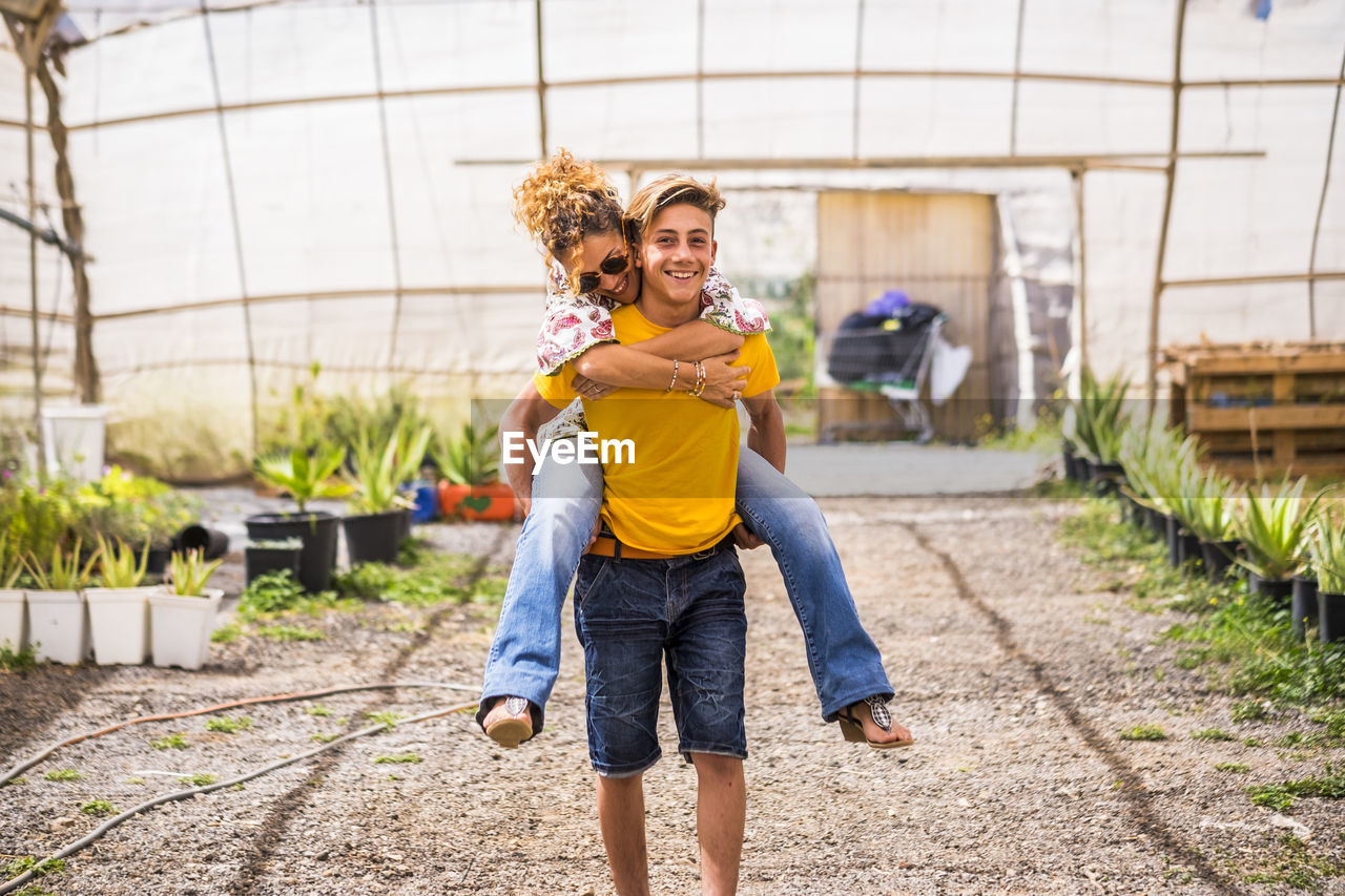 Portrait of teenage boy piggybacking mother in greenhouse