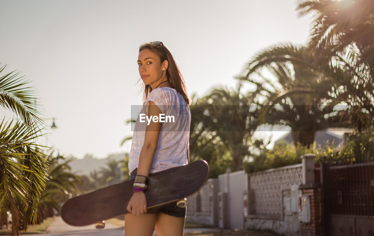 Back view of beautiful young girl with short shorts and skateboard outdoors on a hot summer day.