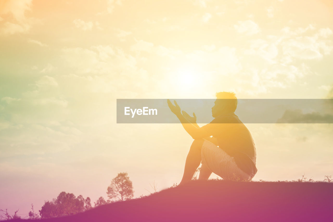 Silhouette man praying while sitting on land against sky during sunset