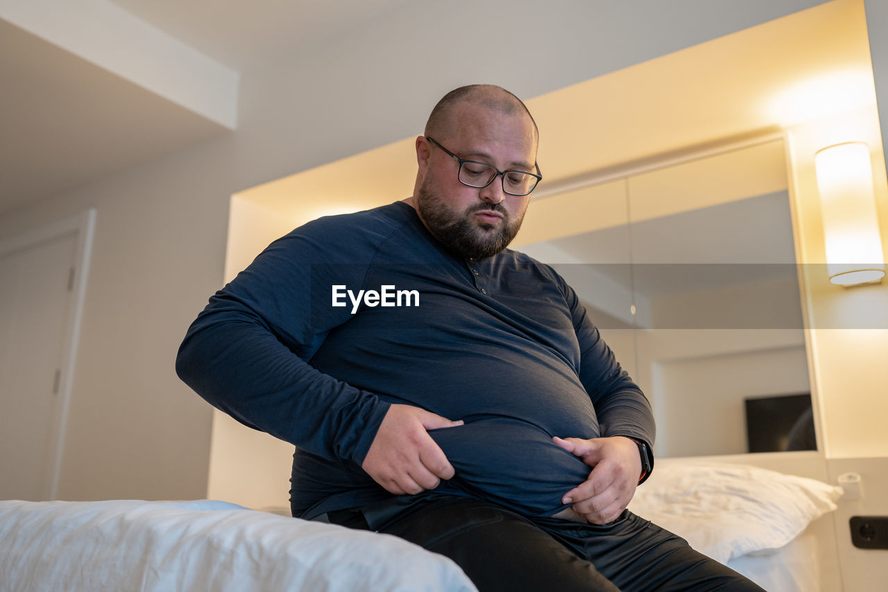 Sad fat man sitting at home bed holds stomach have health problem, insecurities, difficult relations