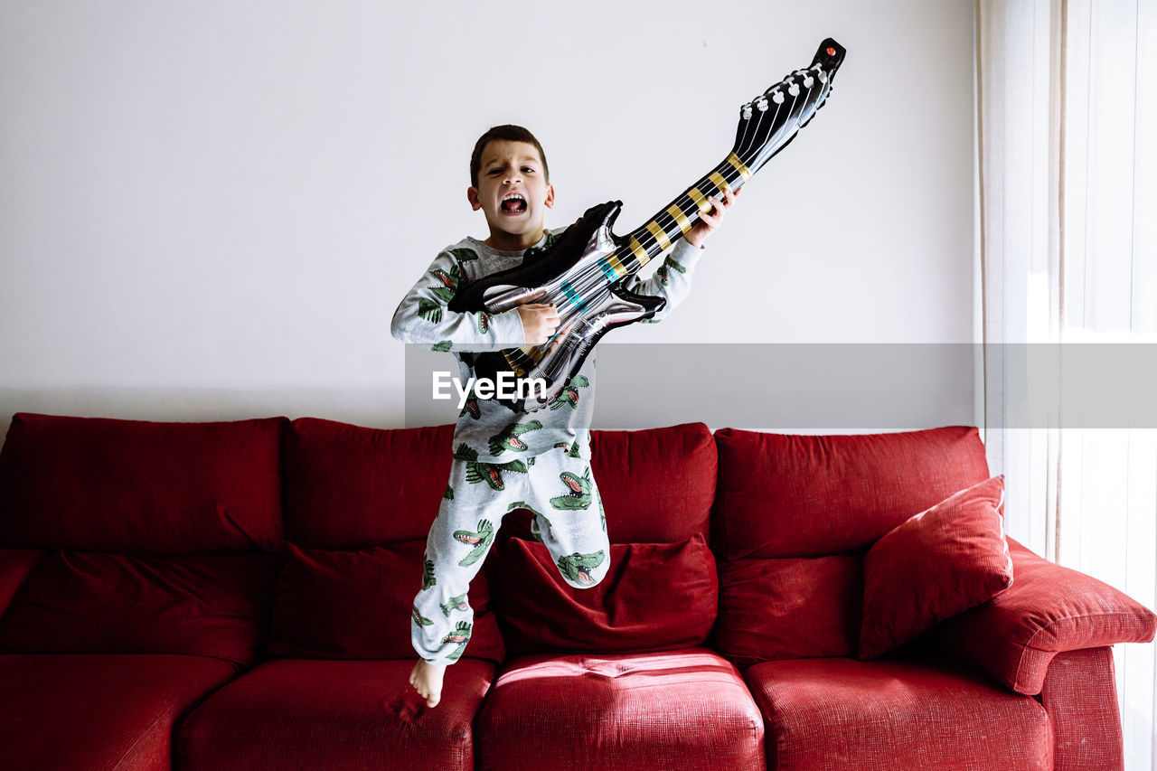 Boy jumping from sofa while playing guitar in living room