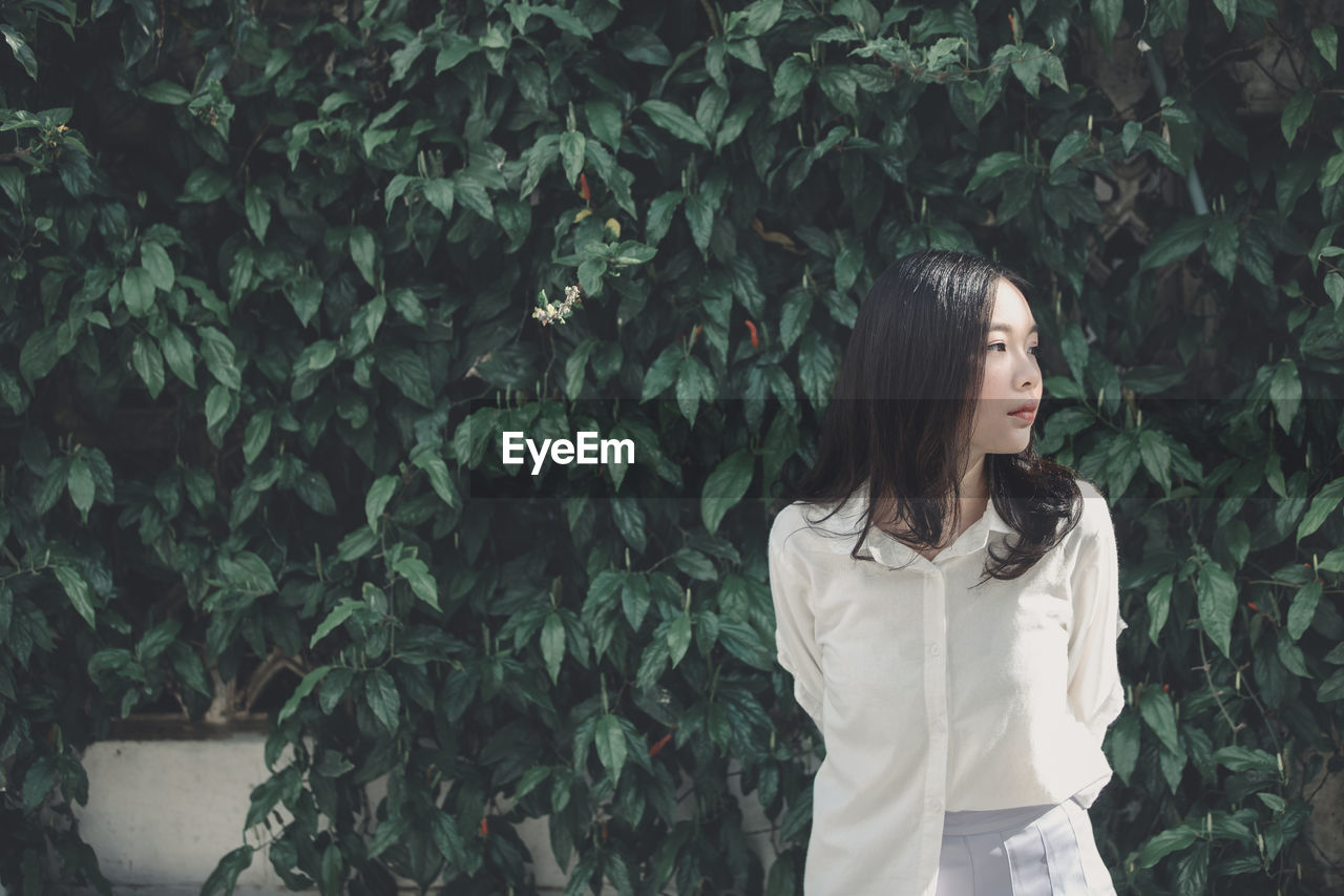 Young woman looking away while standing against plants