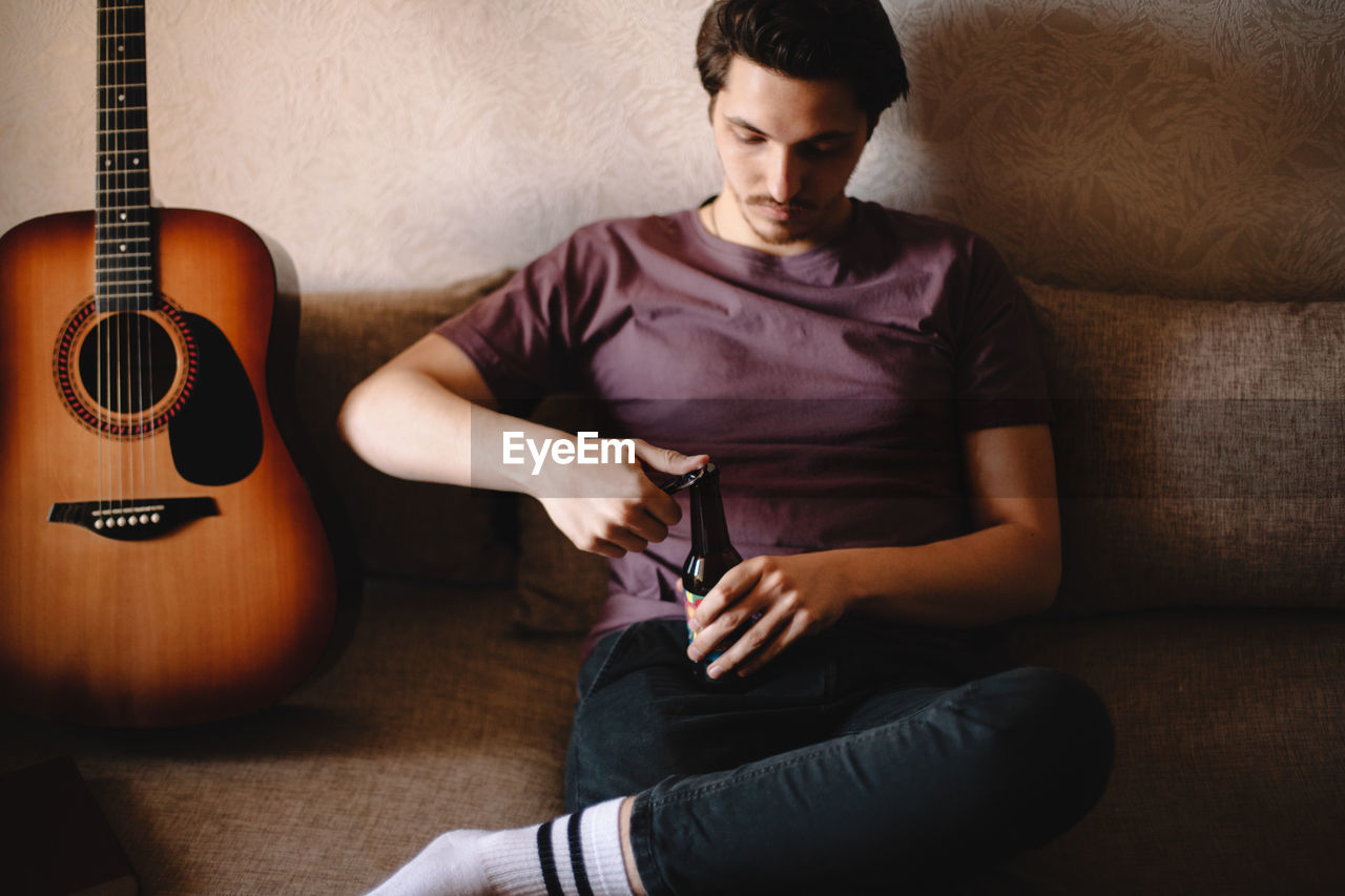 Young man opening bottle of beer sitting on sofa at home