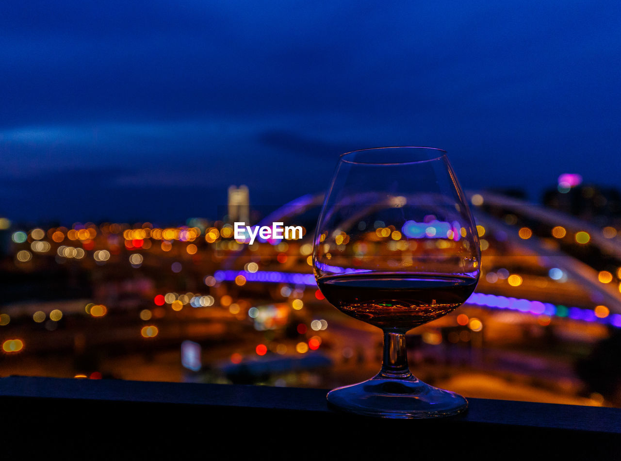 Close-up of wineglass on railing by illuminated cityscape against sky at night