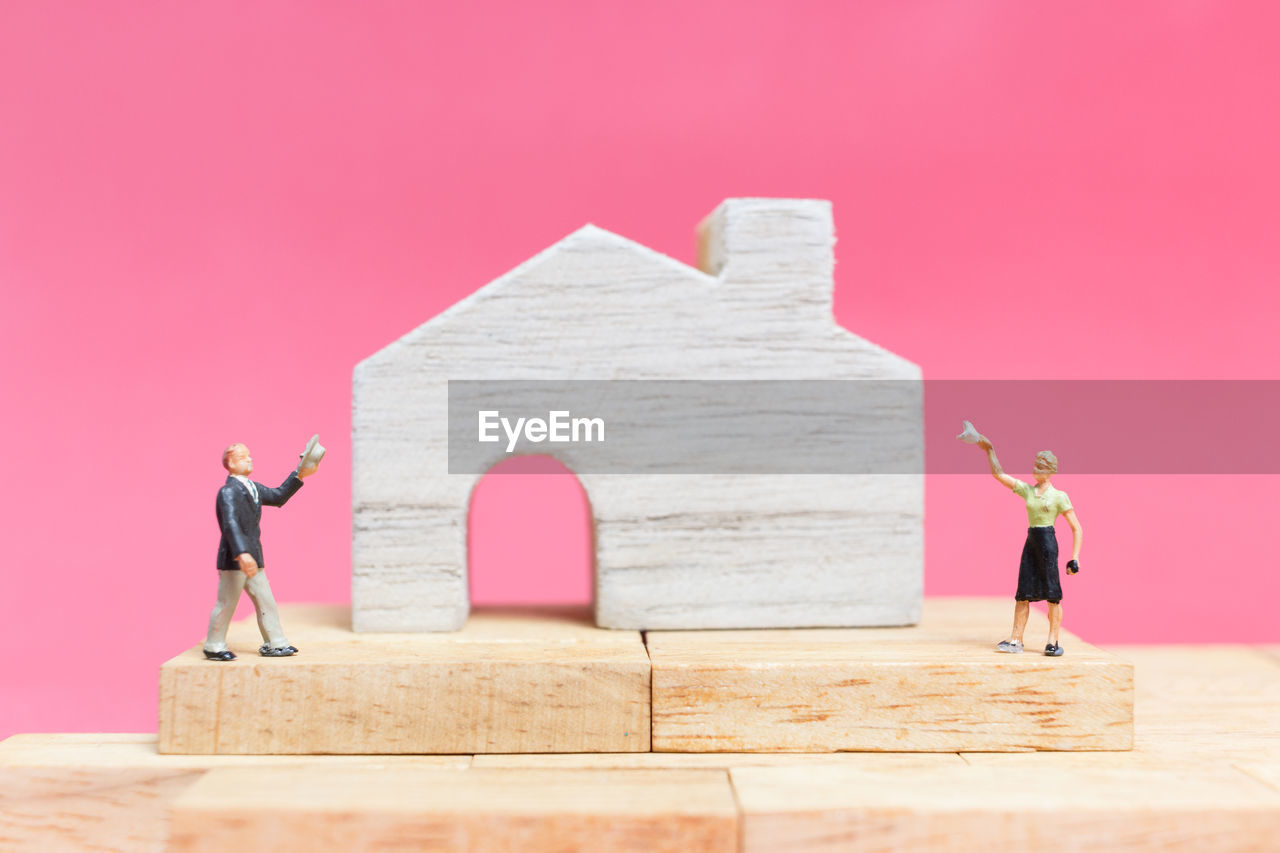 Close-up of figurines with model home on table against pink background