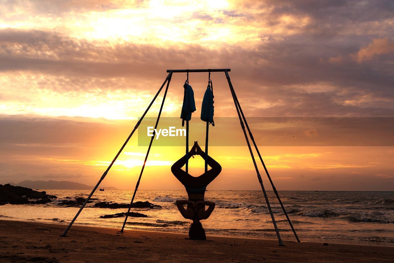 Man doing yoga at beach against sky during sunset