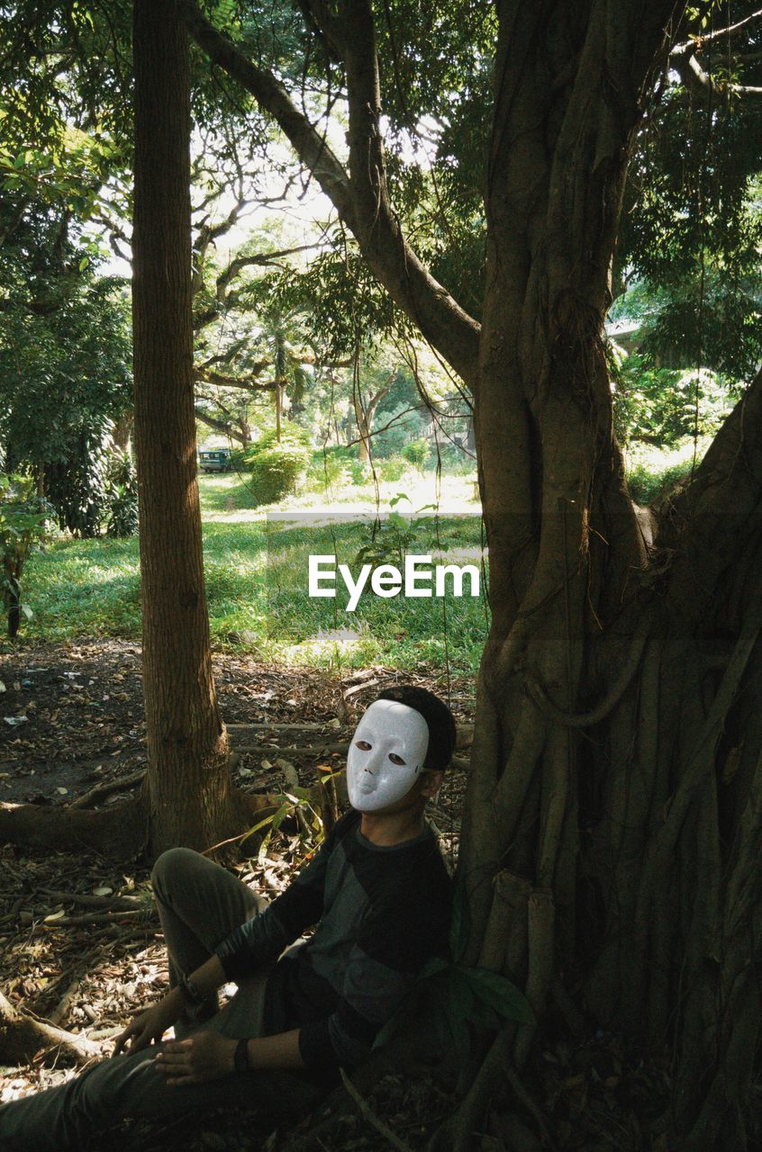 Man wearing mask while sitting by tree on field