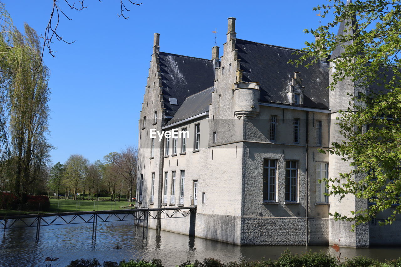 architecture, château, built structure, building exterior, building, tree, plant, nature, sky, estate, water, history, water castle, the past, blue, clear sky, no people, travel destinations, castle, house, moat, outdoors, day, sunny, tourism, mansion, residential district, travel, old, home, stately home, waterway, lake, tower