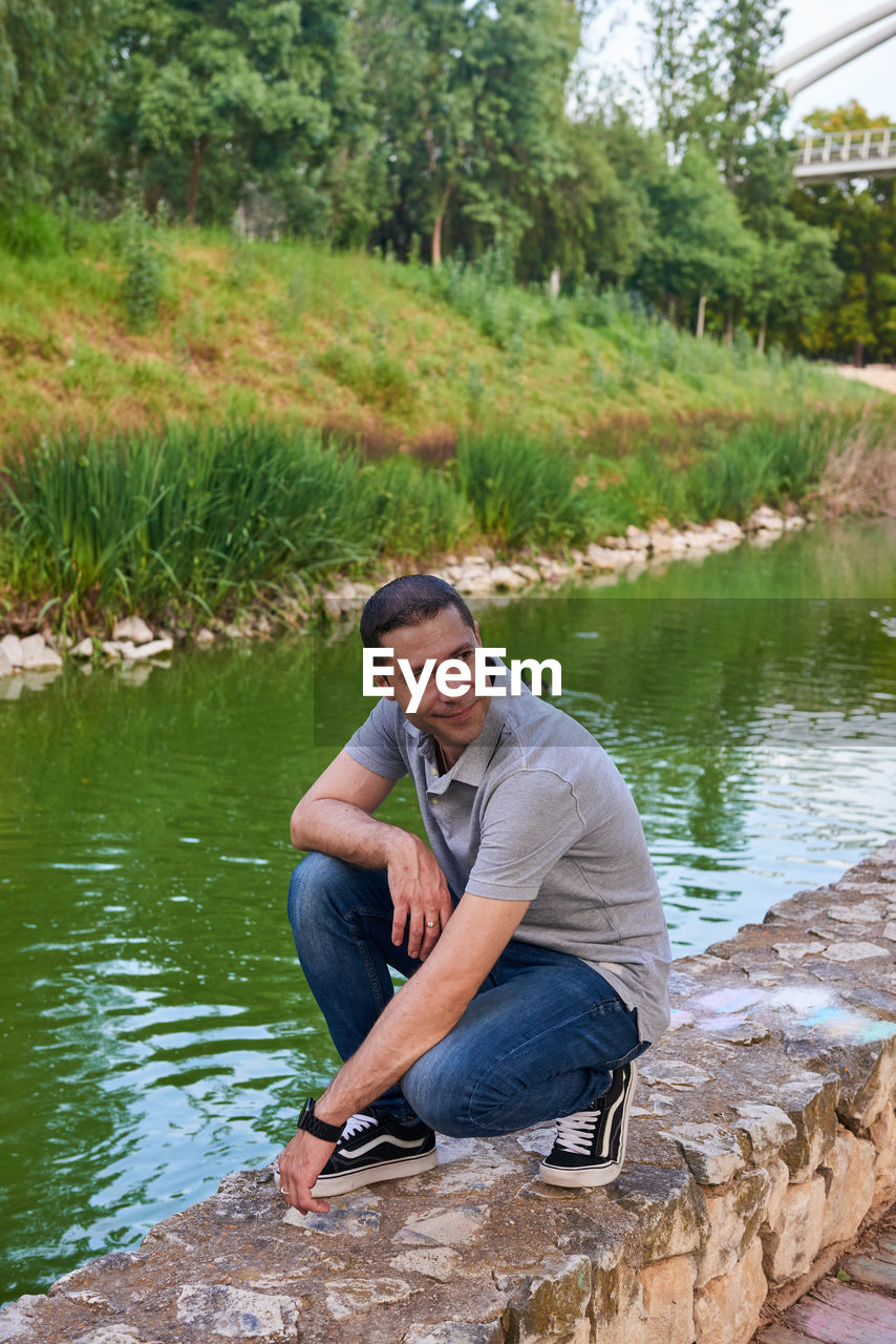 water, one person, sitting, adult, full length, men, nature, plant, lake, leisure activity, casual clothing, tree, day, lifestyles, young adult, outdoors, rock, looking, relaxation, beauty in nature, smiling, tranquility, holiday, crouching, activity, vacation, emotion