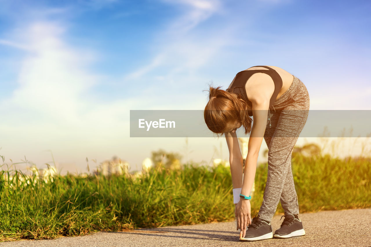 Young woman stretching while standing on road against sky