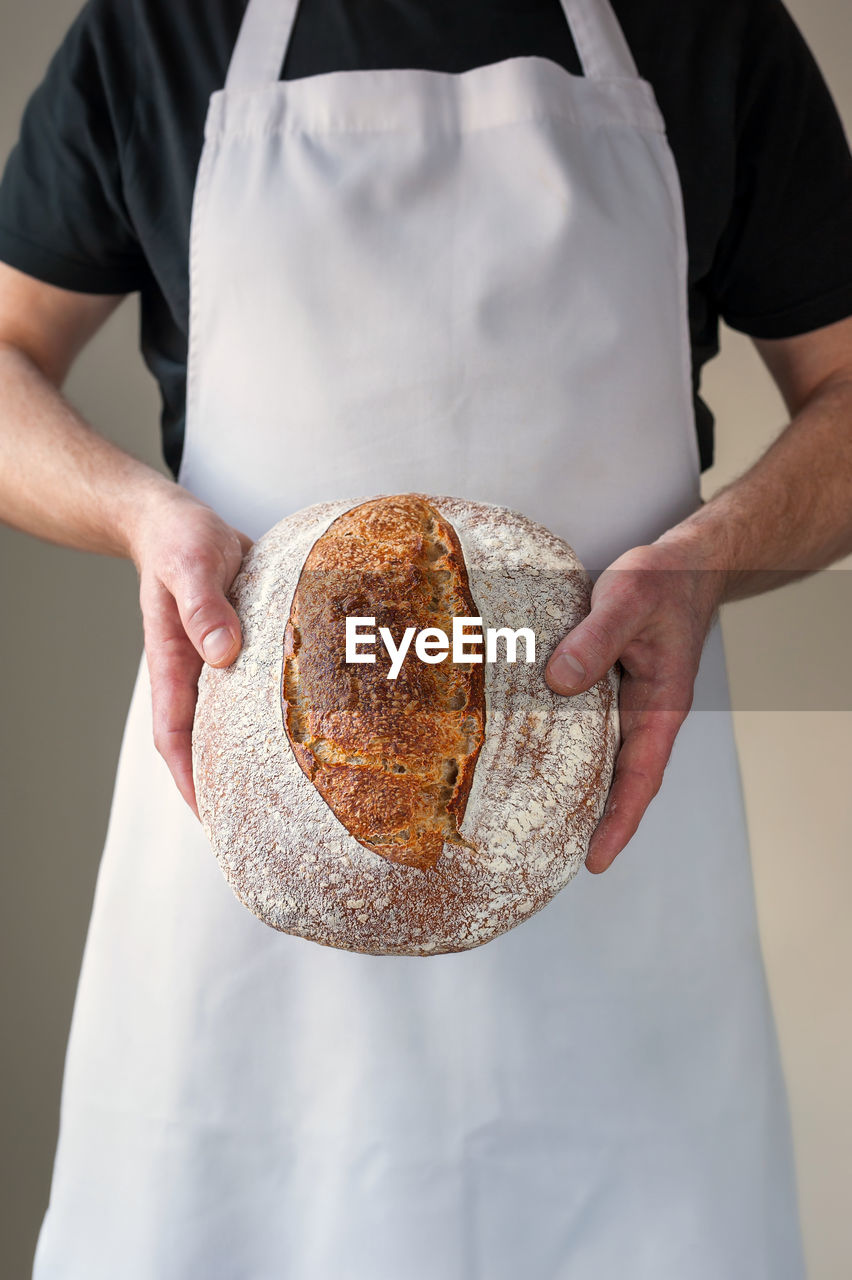 Midsection, bakers hands holding a round shaped loaf of organic sourdough bread in front of him.