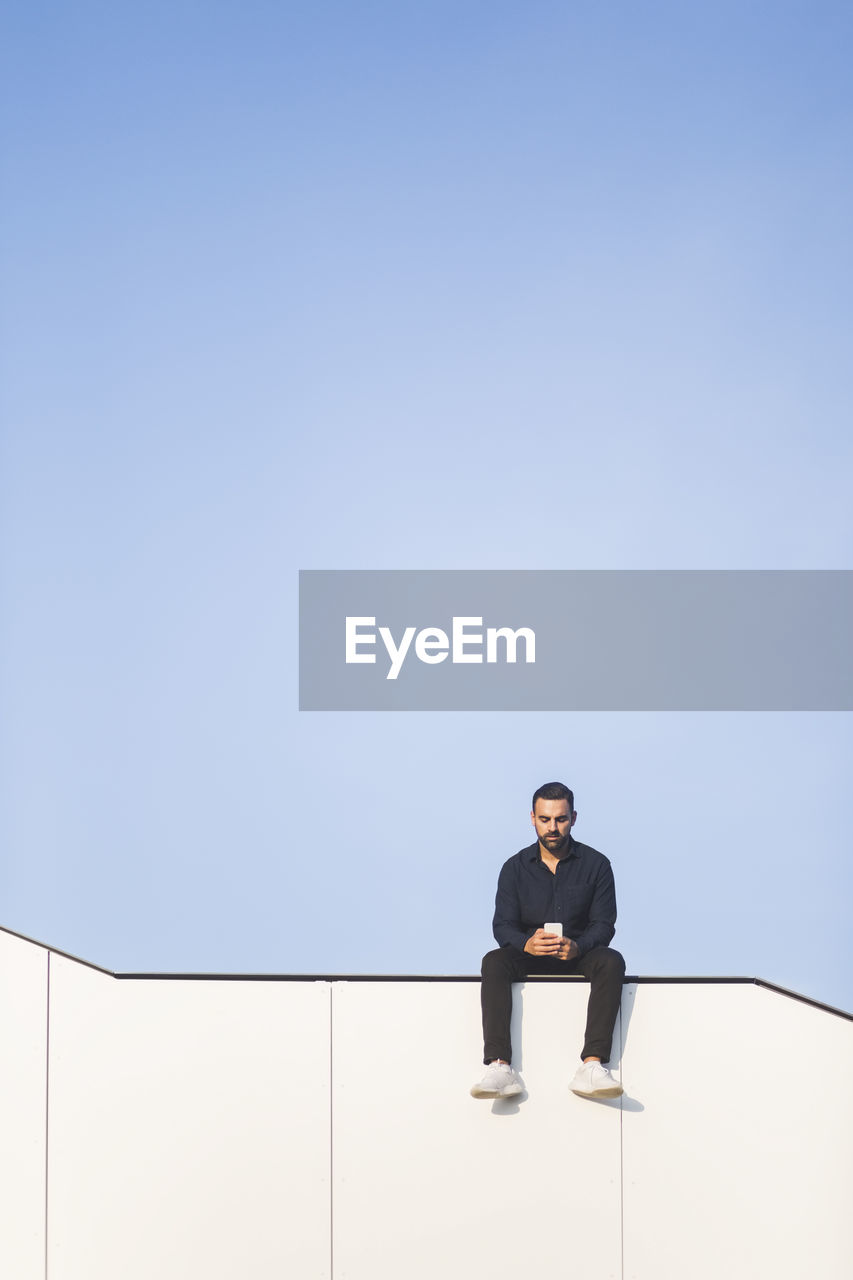 Man using phone while sitting on built structure against blue sky