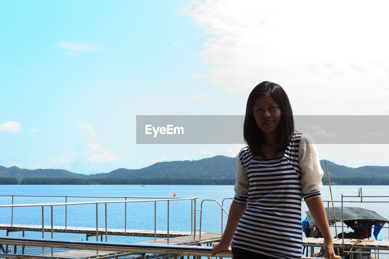 YOUNG WOMAN STANDING BY RAILING AGAINST SKY WITH WATER