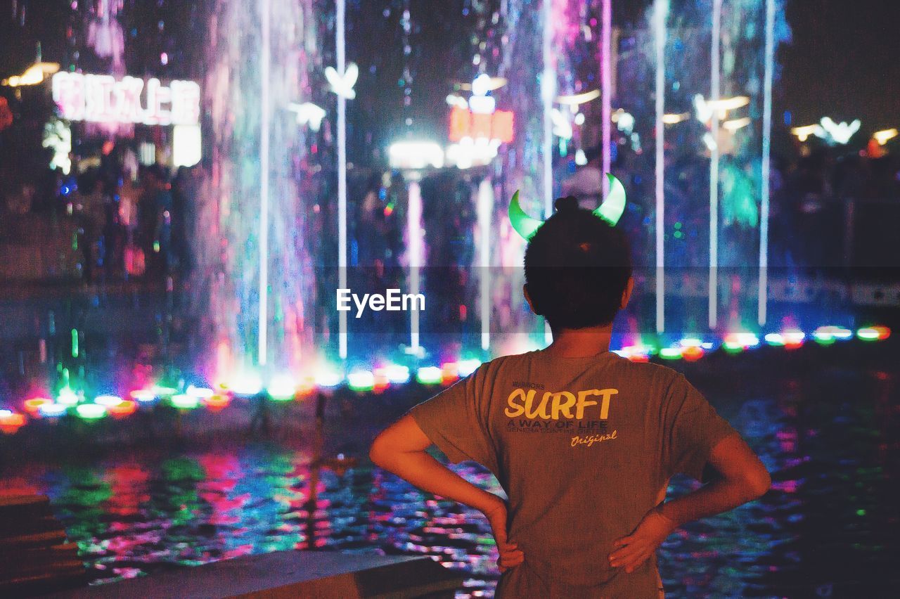 Rear view of boy viewing illuminated fountain at night in amusement park