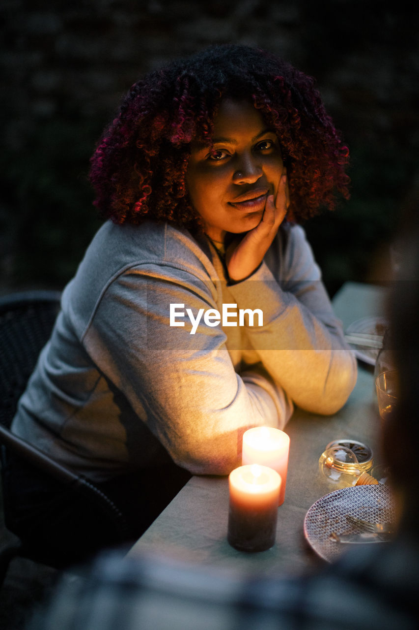 Portrait of young woman with curly hair by illuminated candle on dining table