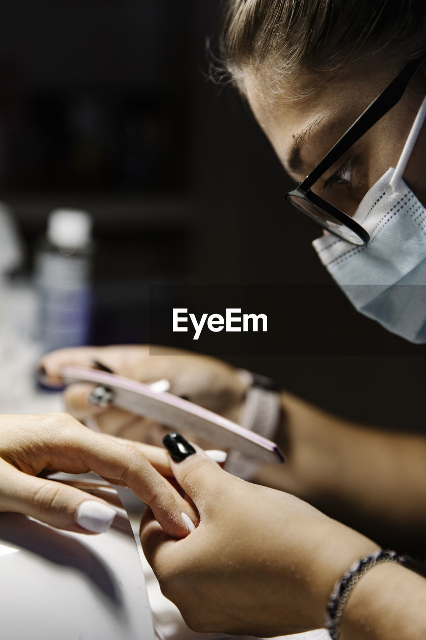 Woman in mask shaping nails of client while working during epidemic