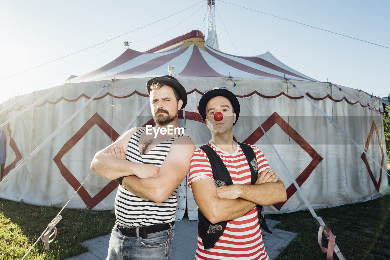 Muscular build artist standing with arms crossed by clown in front of circus tent