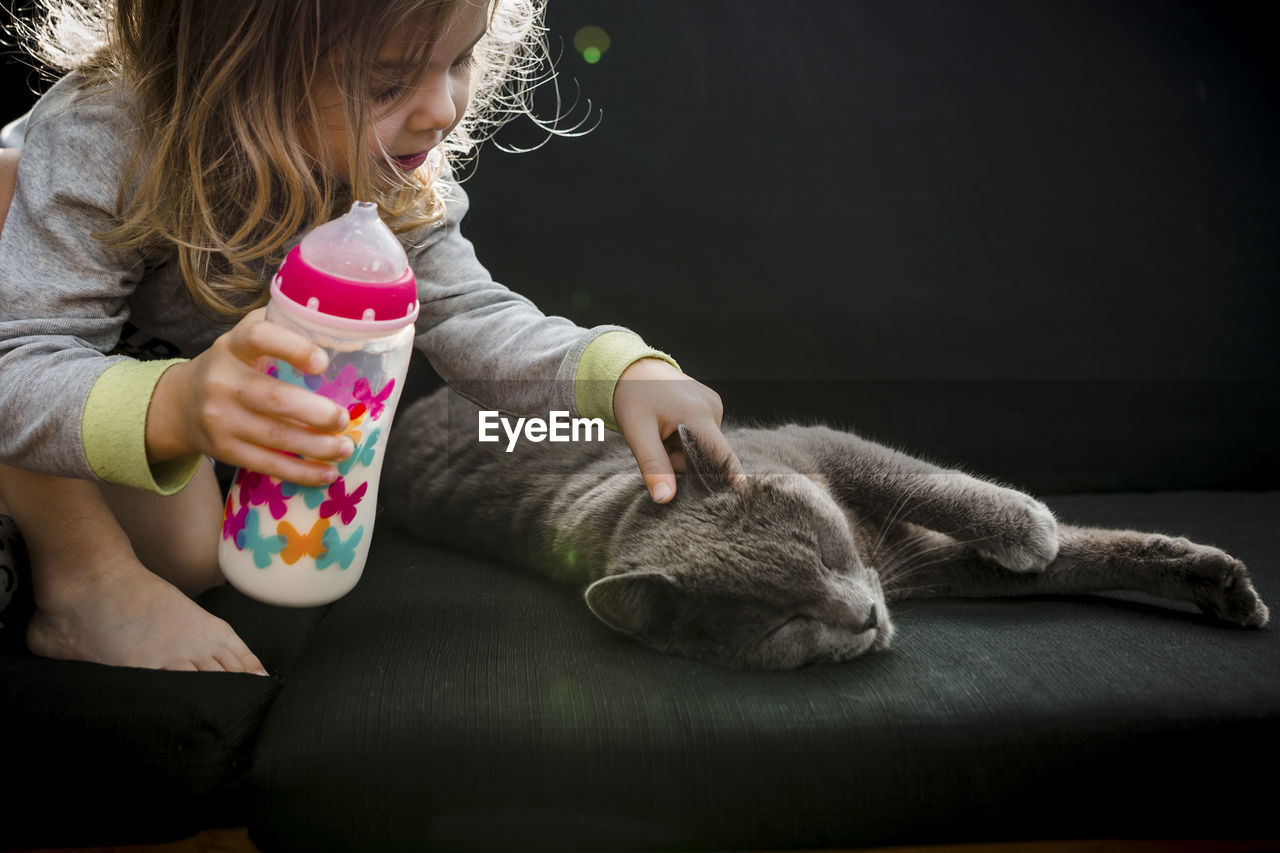 Toddler girl patting her grey cat pet while drinking milk outdoor