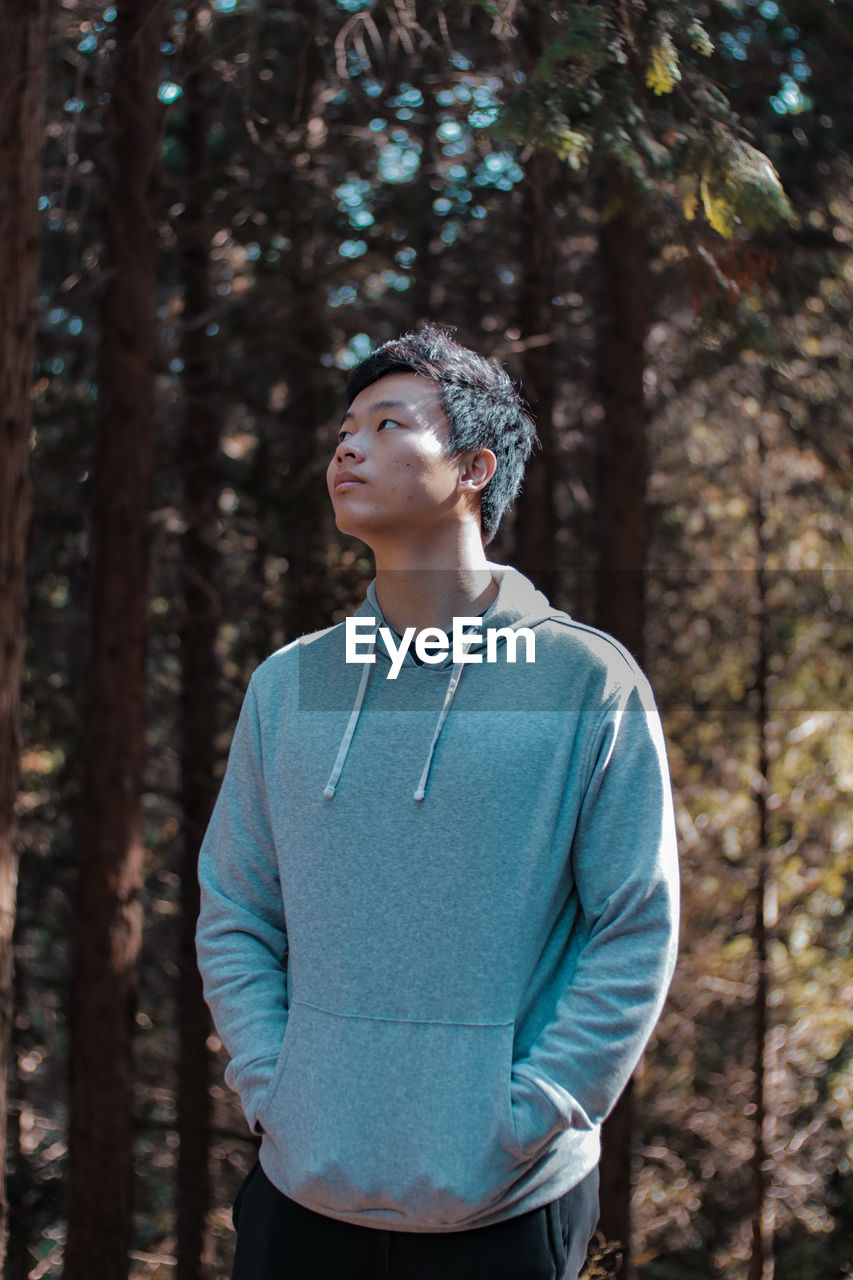 Young man looking away while standing against trees