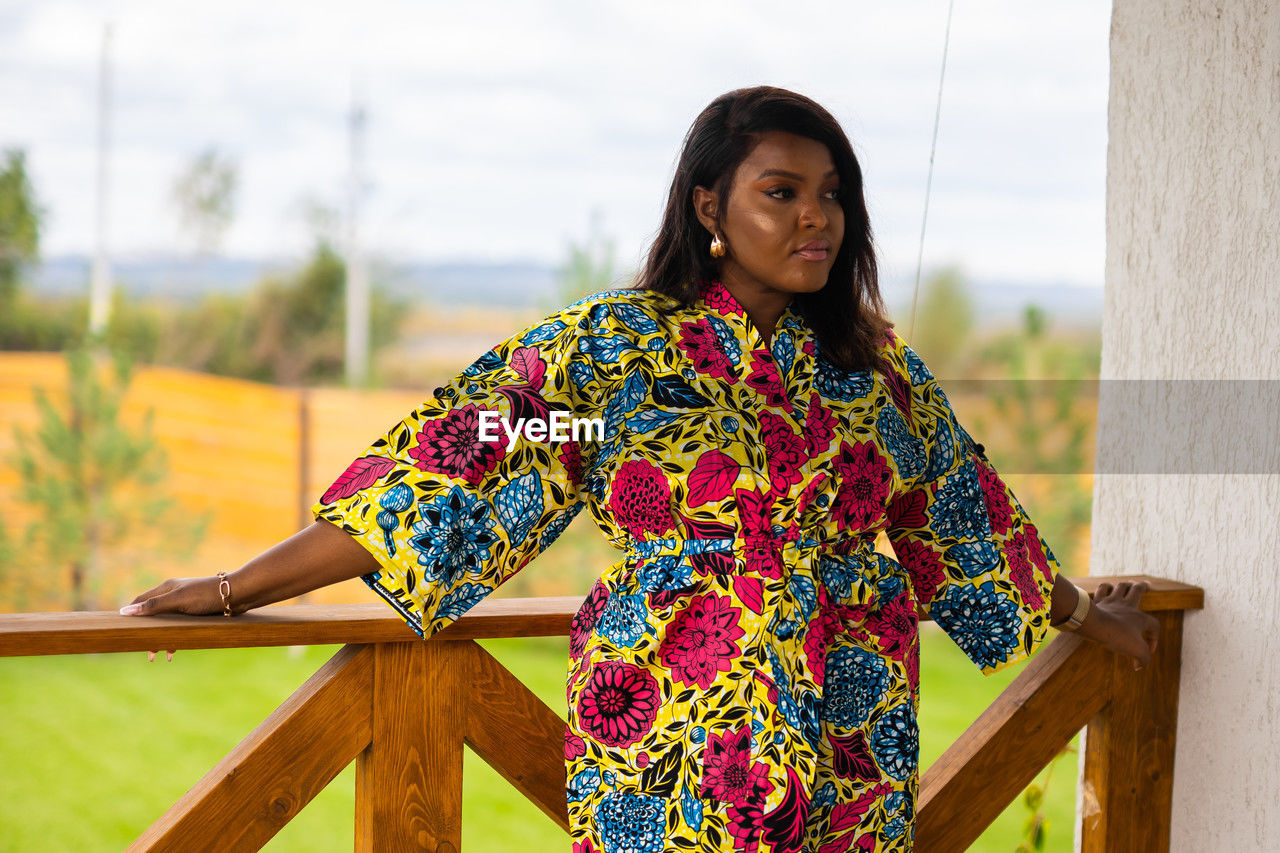 one person, spring, yellow, women, adult, dress, standing, portrait, nature, clothing, three quarter length, photo shoot, smiling, lifestyles, young adult, hairstyle, floral pattern, leisure activity, pattern, waist up, outdoors, looking at camera, day, relaxation, female, front view, happiness, multi colored, focus on foreground, architecture, fashion, sky, looking, emotion, casual clothing, wood, black hair, traditional clothing, person, landscape, long hair