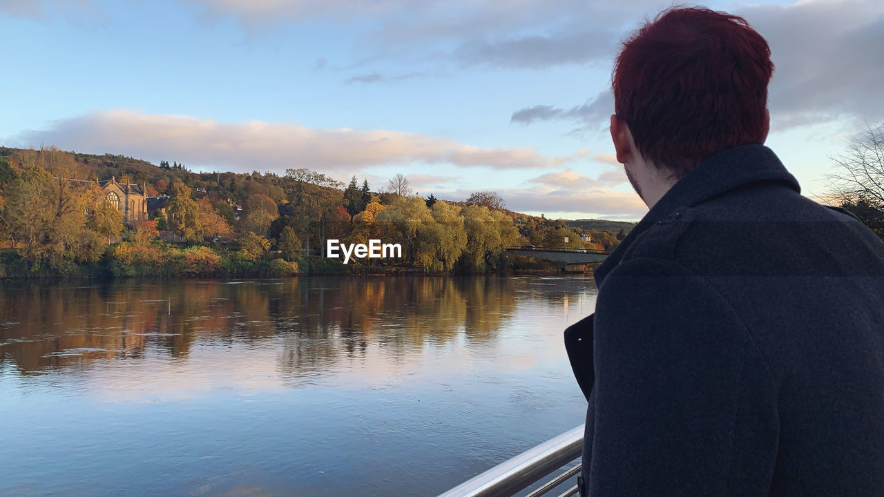 REAR VIEW OF MAN STANDING BY LAKE AGAINST SKY