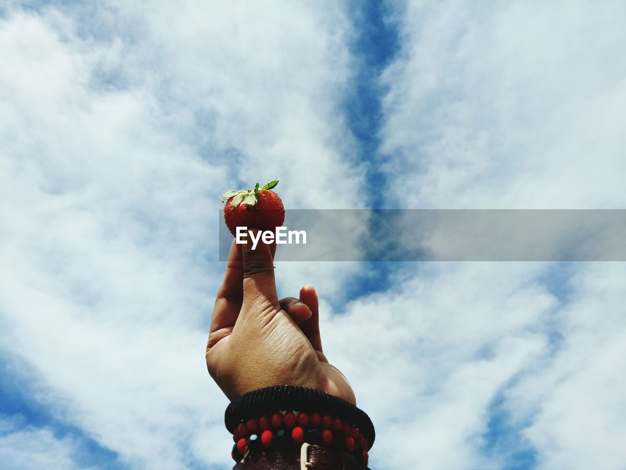 Cropped hand holding strawberry against cloudy sky