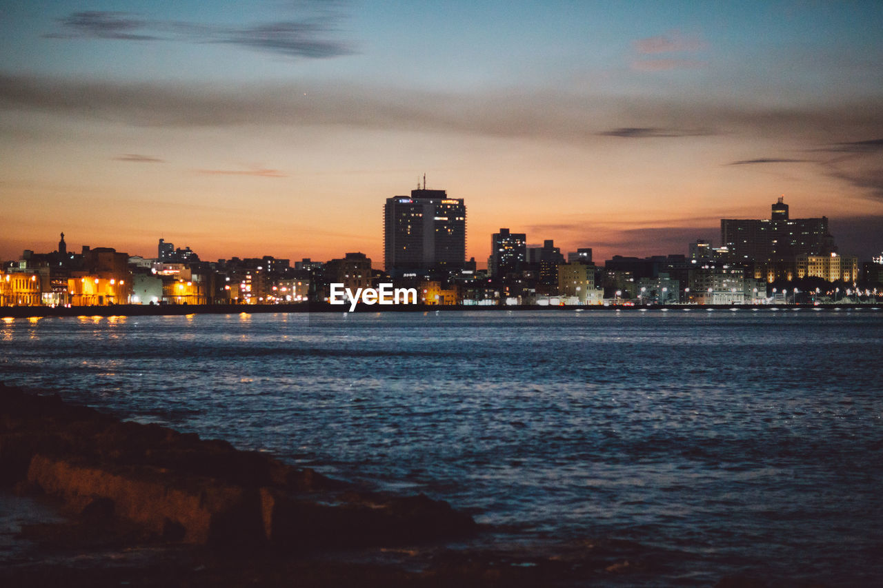 Illuminated city by sea against sky at dusk