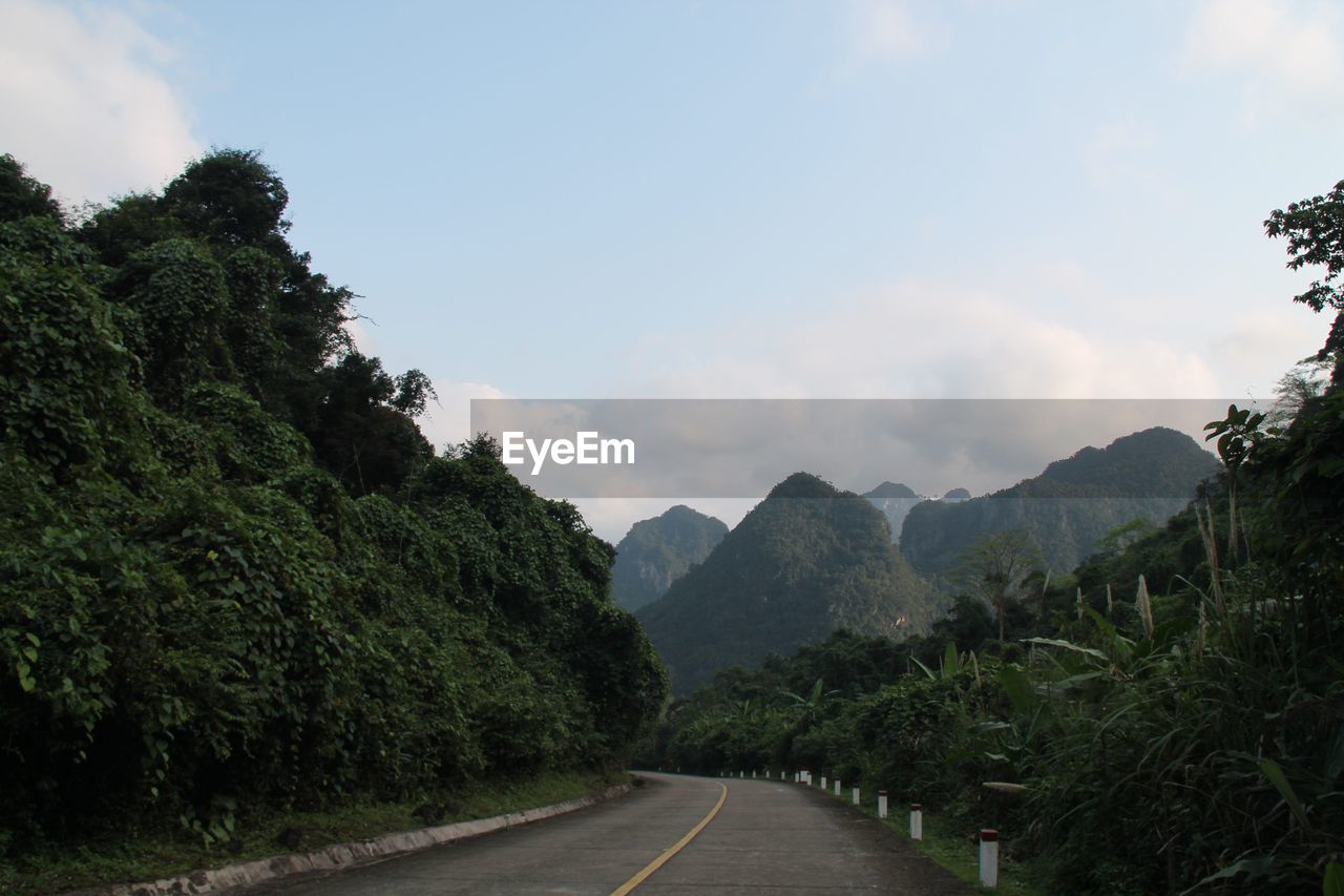 Empty road along trees and mountains against sky