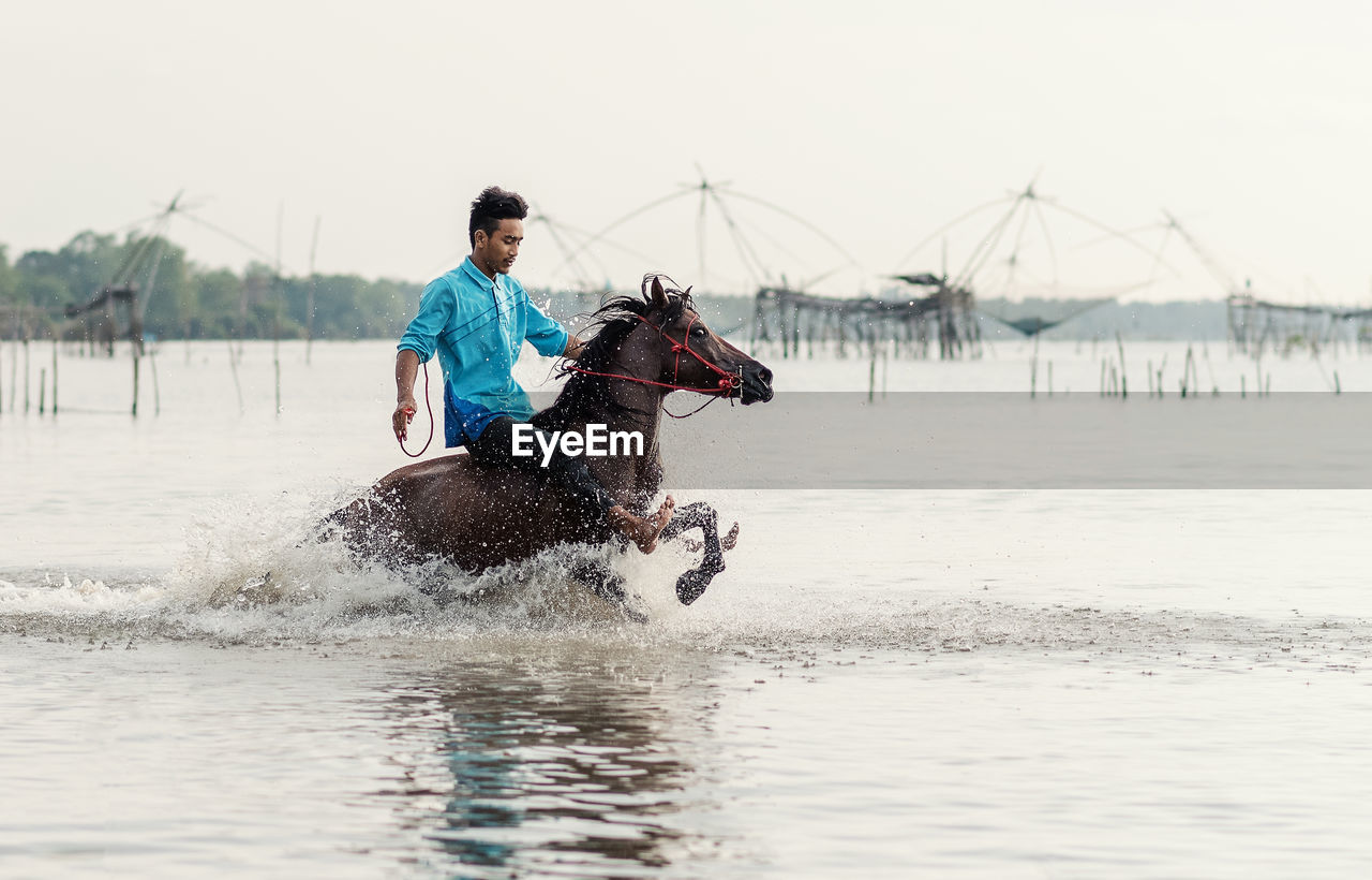 Man riding horse in lake against sky during sunset