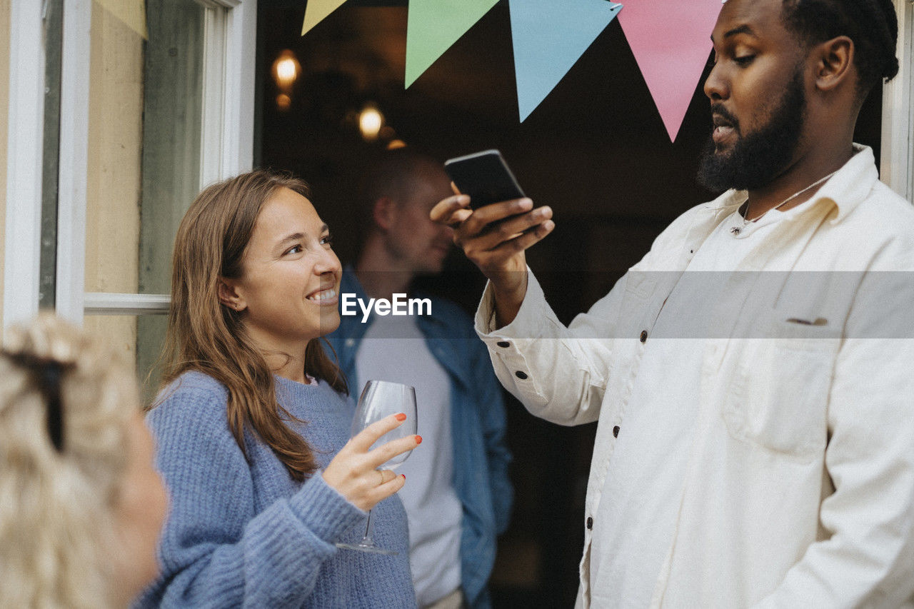 Man taking picture of female friend holding wineglass during dinner party at cafe