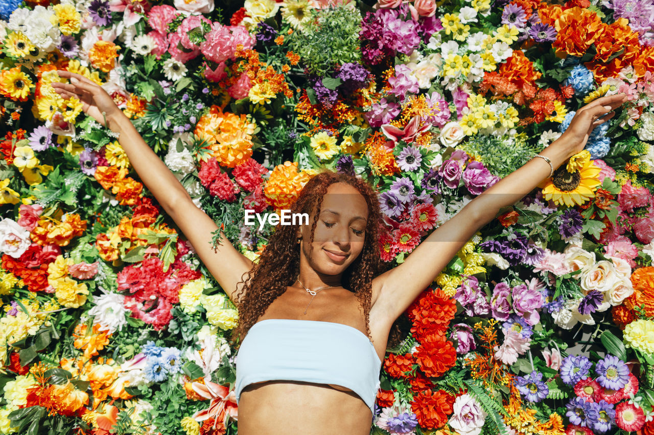 Smiling young woman with arms raised standing in front of flower wall on sunny day