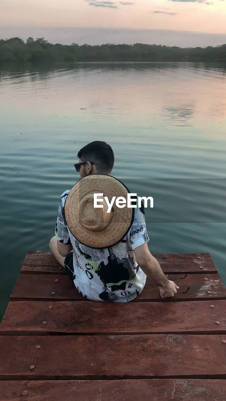 Rear view of man sitting on pier over lake against sky