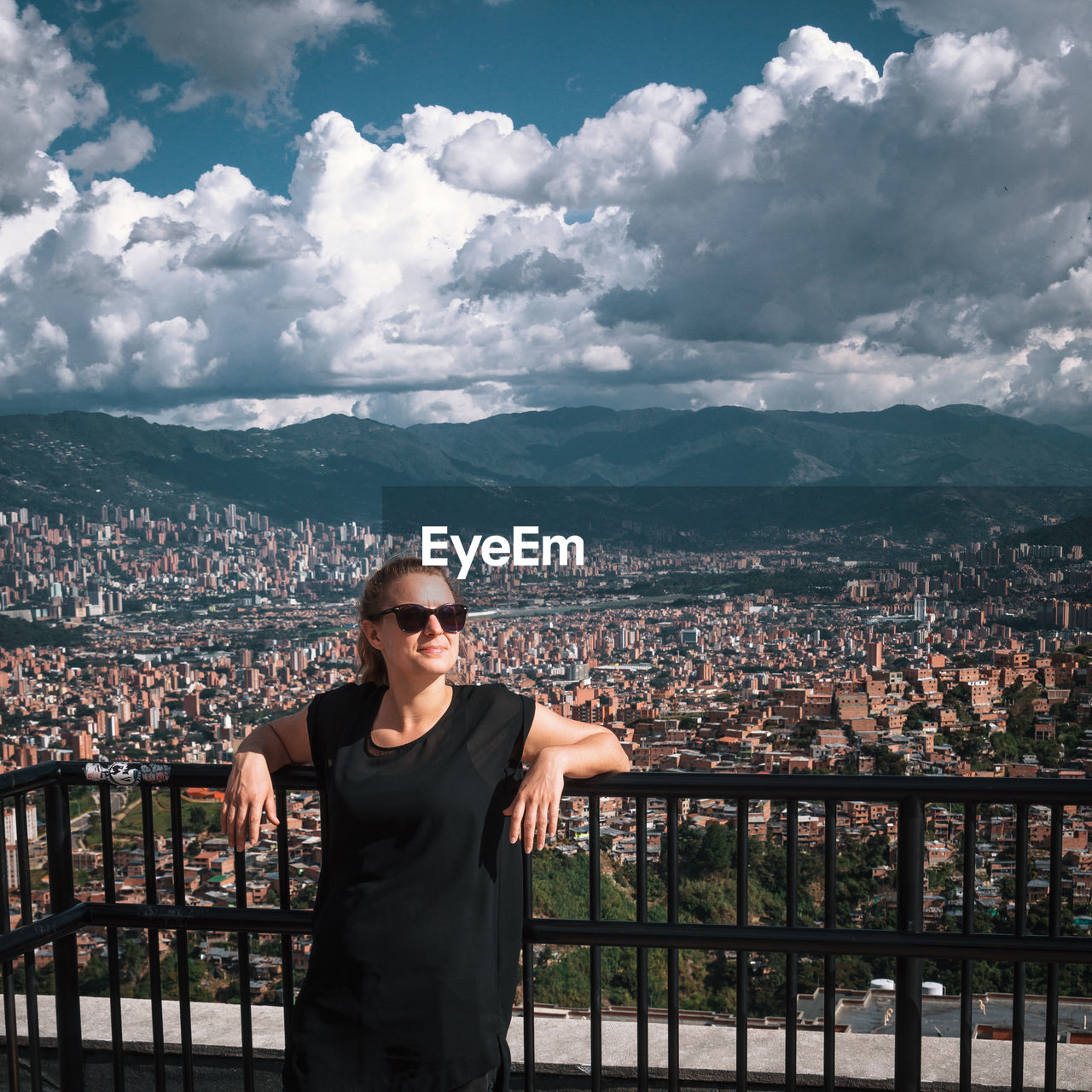 Woman standing at building balcony with city in background against cloudy sky