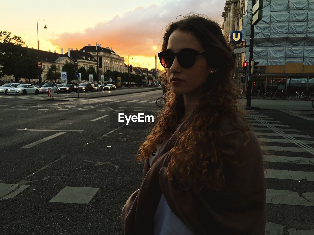 Beautiful young woman in sunglasses standing on street at sunset