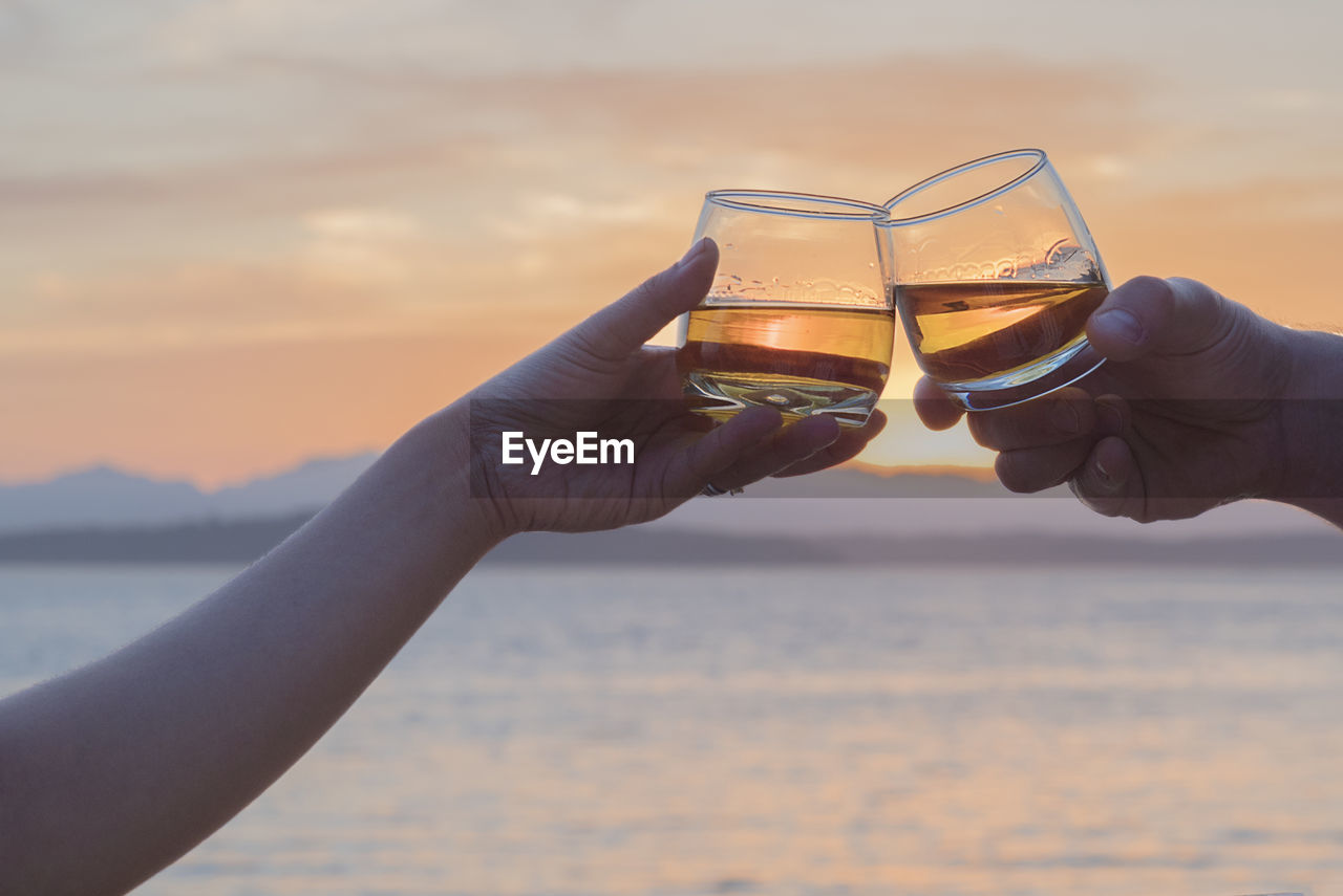 Cropped hands of couple toasting whiskey against sky during sunset