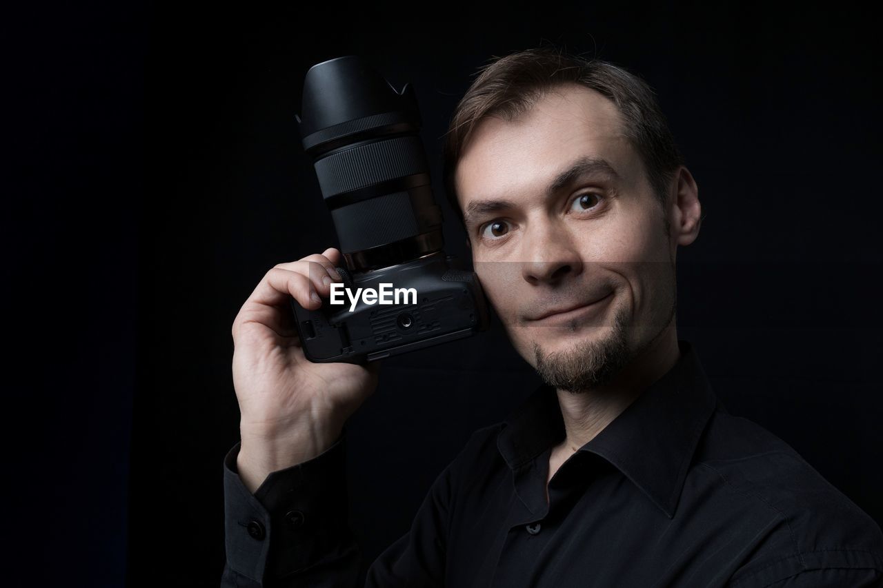 portrait, one person, adult, men, black background, studio shot, looking at camera, technology, person, indoors, headshot, human face, holding, black, camera, smiling, singing, occupation, dark, facial hair, professional occupation, beard, communication, front view, activity, clothing, emotion, cut out, happiness, young adult