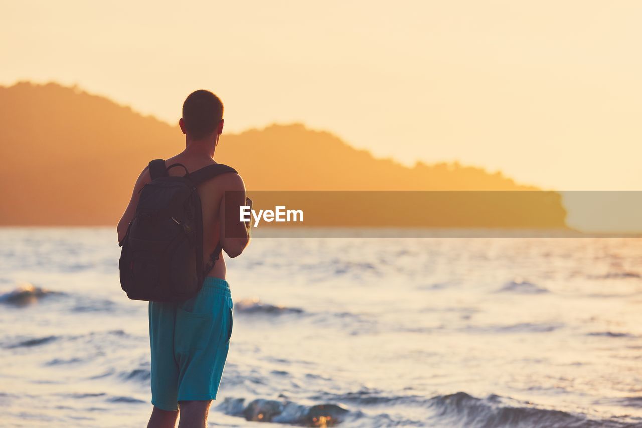 Rear view of man carrying backpack looking at sea during sunset