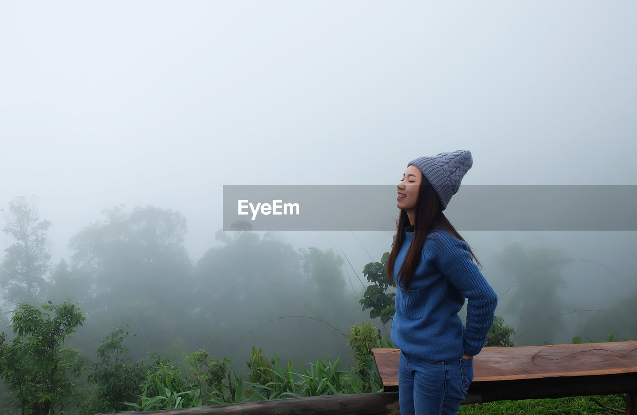 Side view of young woman standing by railing against trees during foggy weather