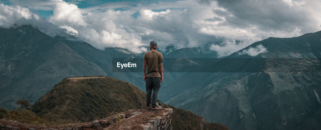 Rear view of young man looking at mountains while standing against cloudy sky