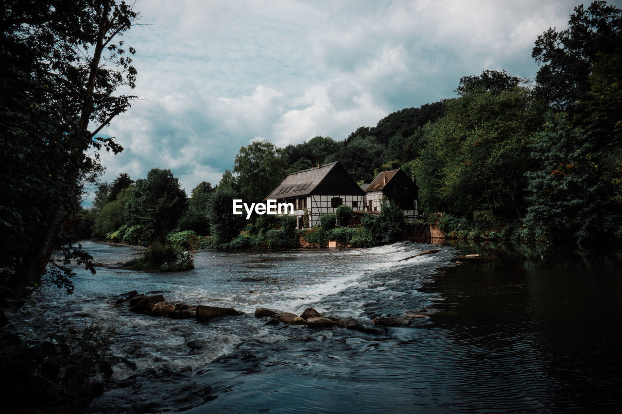 HOUSES BY RIVER AMIDST TREES AND BUILDING AGAINST SKY