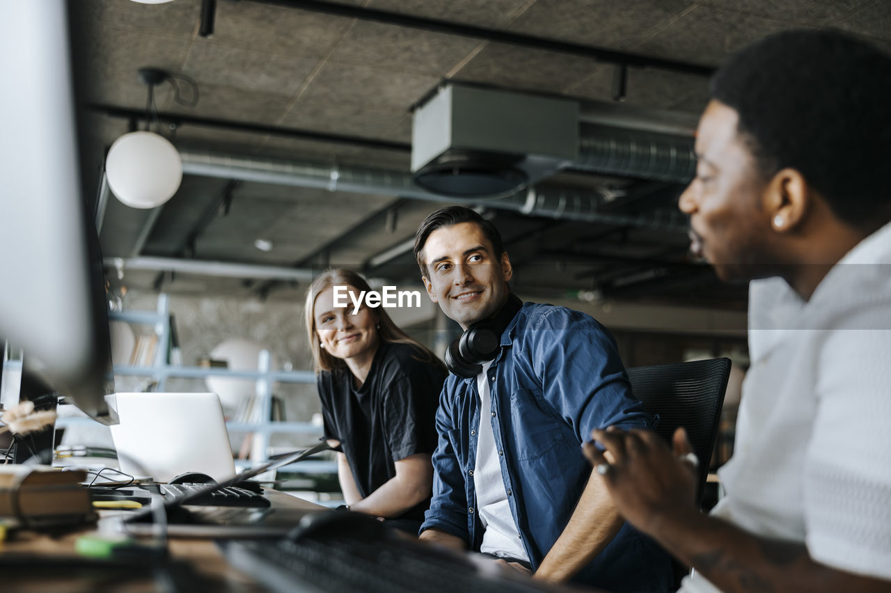 Male and female computer programmers talking with colleague sitting in office